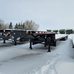 A 2025 Behnke STX 53-foot semi-style sprayer trailer. The trailer is a long, low-profile vehicle designed to transport large agricultural sprayers. It is black and has a unique shape to accommodate the wide sprayer booms.